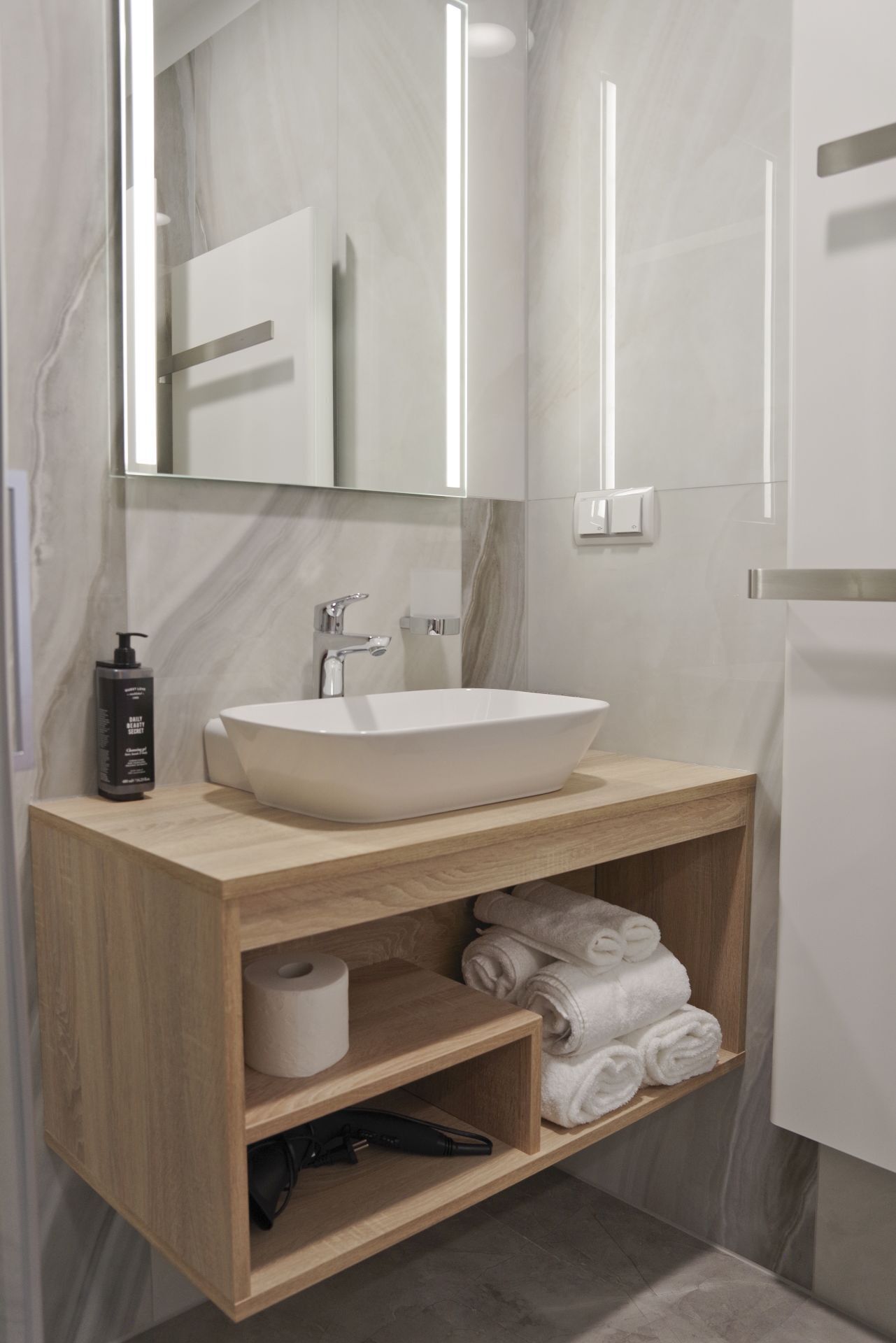 Un baño moderno con un espejo grande, un lavamanos sobre encimera y un mueble de tocador de madera con estantes para toallas y artículos de aseo.