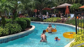 A swimming pool area with lounge chairs and umbrellas, surrounded by greenery and a building in the background.