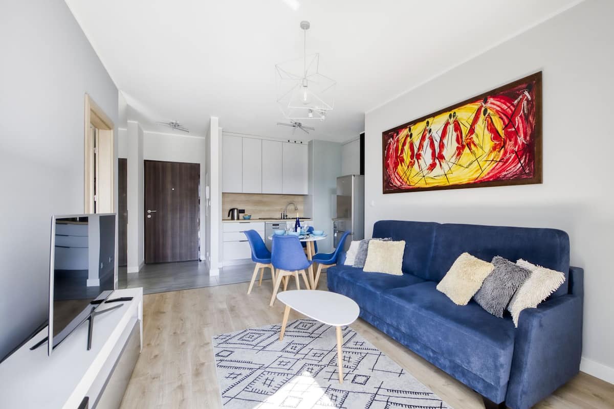 A modern living room with a blue sofa, a dining area with blue chairs, a kitchen in the background, and a large abstract painting on the wall.