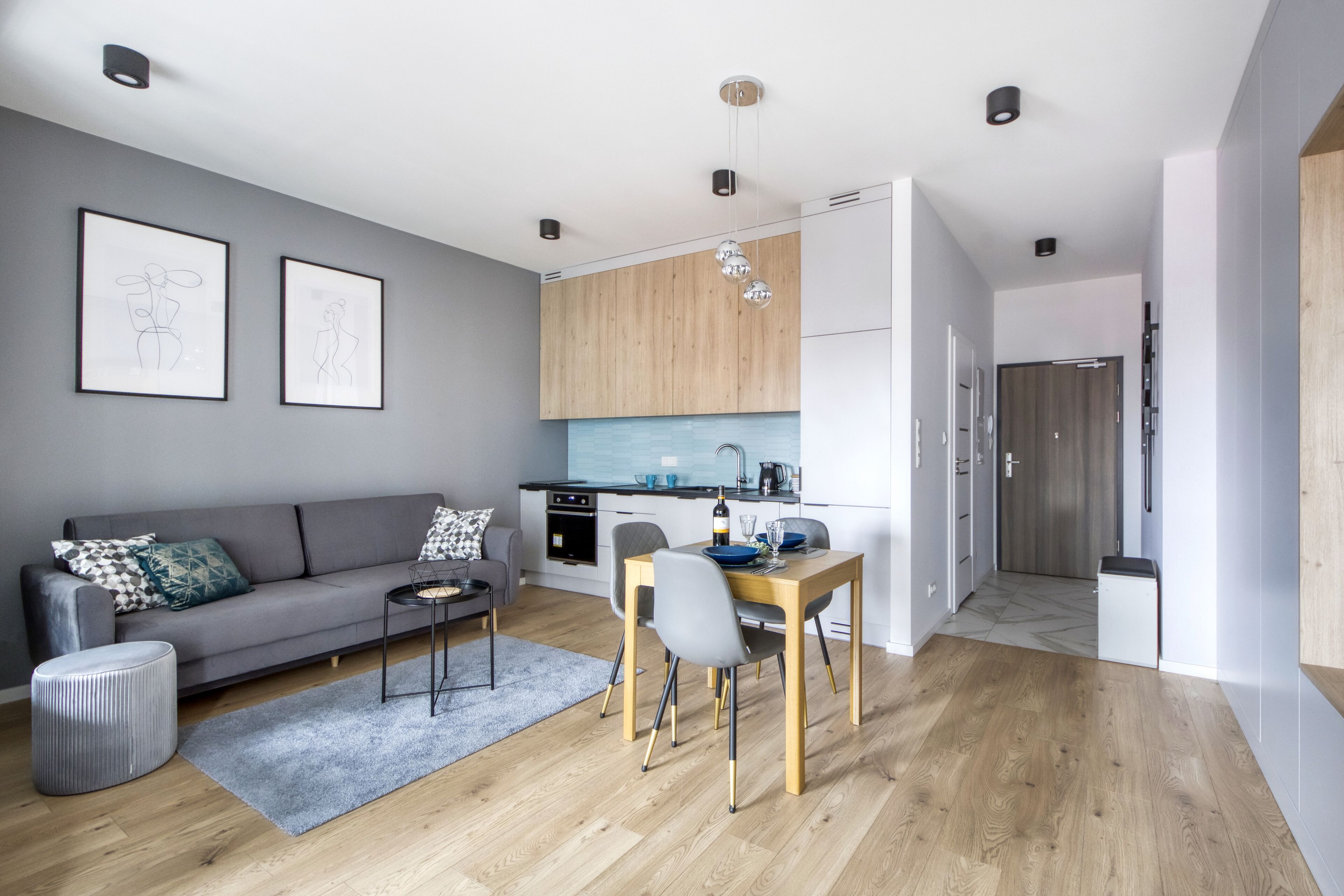 Un salon moderne avec un canapé gris, une table à manger avec des chaises et un coin cuisine doté de meubles en bois et d’un plan de travail bleu.