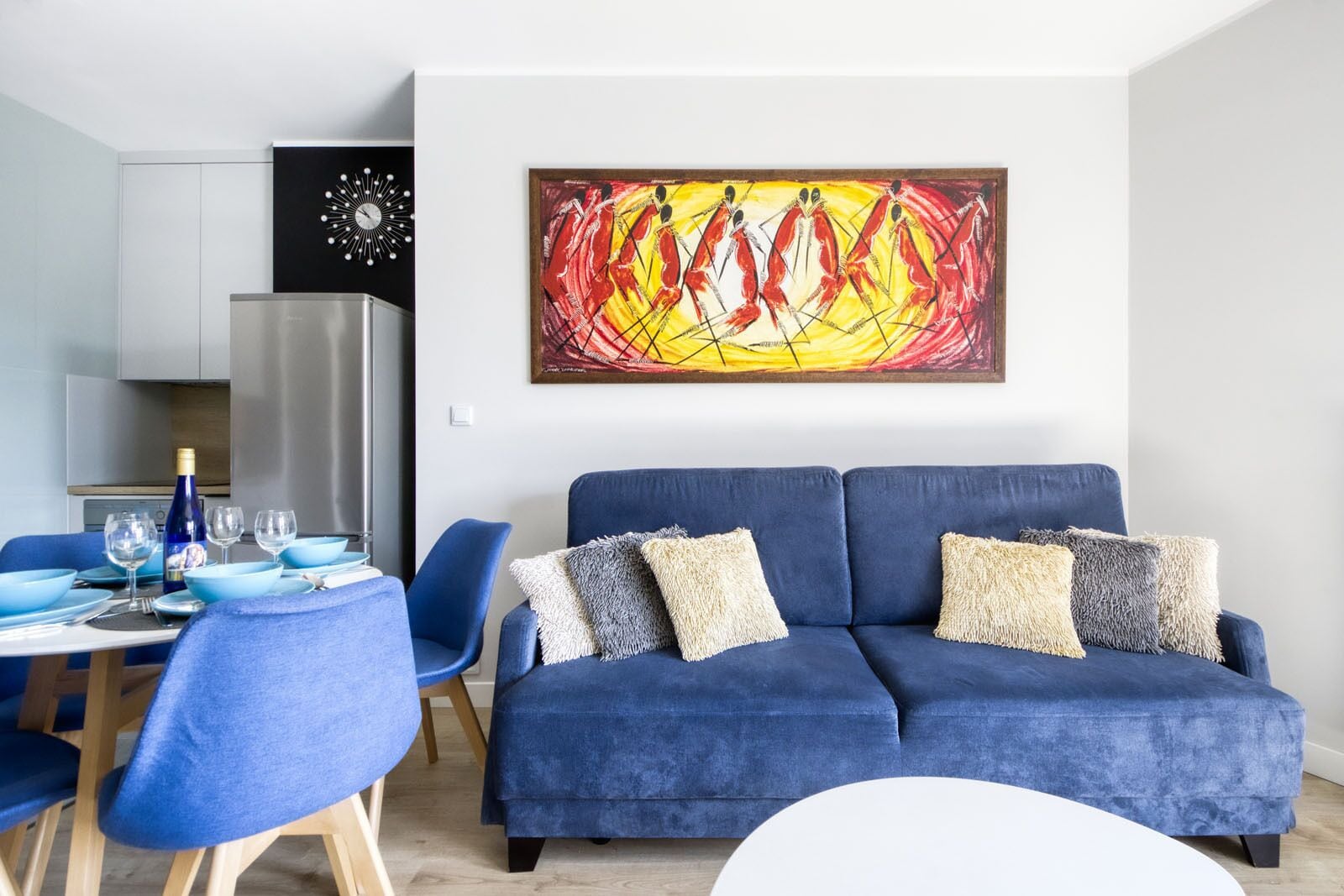Een moderne woonkamer met een blauwe bank, een eethoek met blauwe stoelen en een keuken met een roestvrijstalen koelkast.