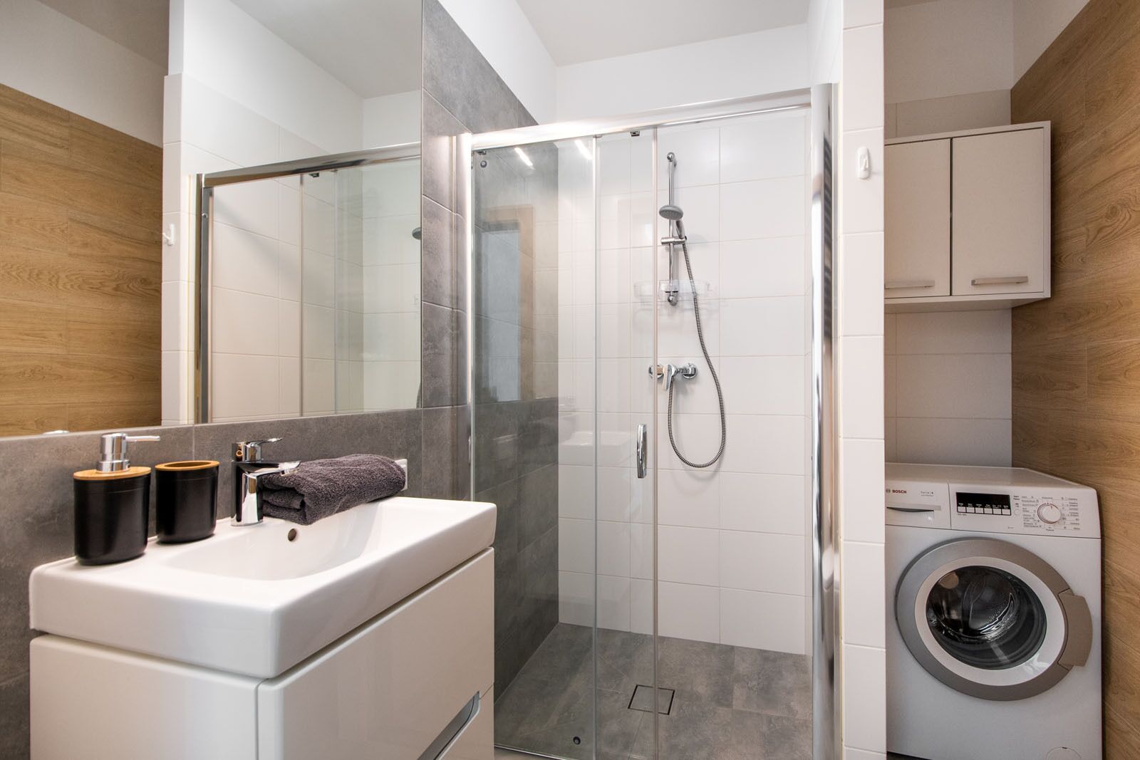 Un baño moderno con ducha de vidrio, lavadora integrada y tocador con lavabo y dispensador de toallas.