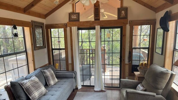 A cozy living room with a gray sofa, a gray armchair, and a wooden floor. There are large windows with curtains, a ceiling fan, and framed pictures on the walls. - Quiet cabin on the Brazos
 (Graham)