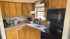 A compact kitchen with wooden cabinets, a black stove, a microwave, and a window providing natural light. - Quiet cabin on the Brazos
 (Graham)