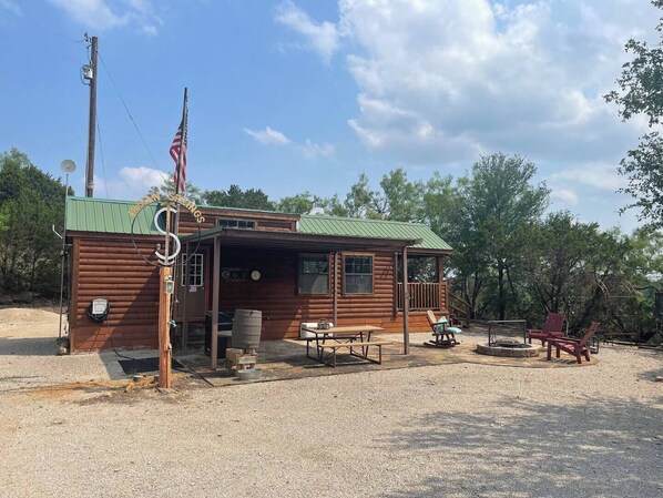 Une cabane rustique avec un toit végétalisé, une véranda avec une table et des chaises, et un drapeau américain.