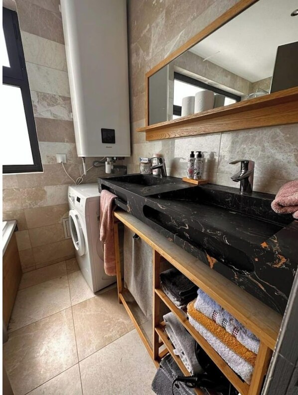 Un baño moderno con lavamanos de mármol negro, lavadora empotrada, tocador de madera con estantes y un espejo grande.