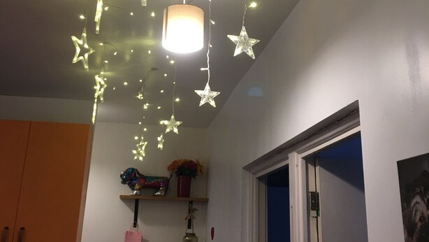 A room with a ceiling light fixture, star-shaped hanging lights, a wooden chair, a shelf with decorative items, and a door with a curtain. - Beautiful 2-bed Apartment in Dagenham (Dagenham)