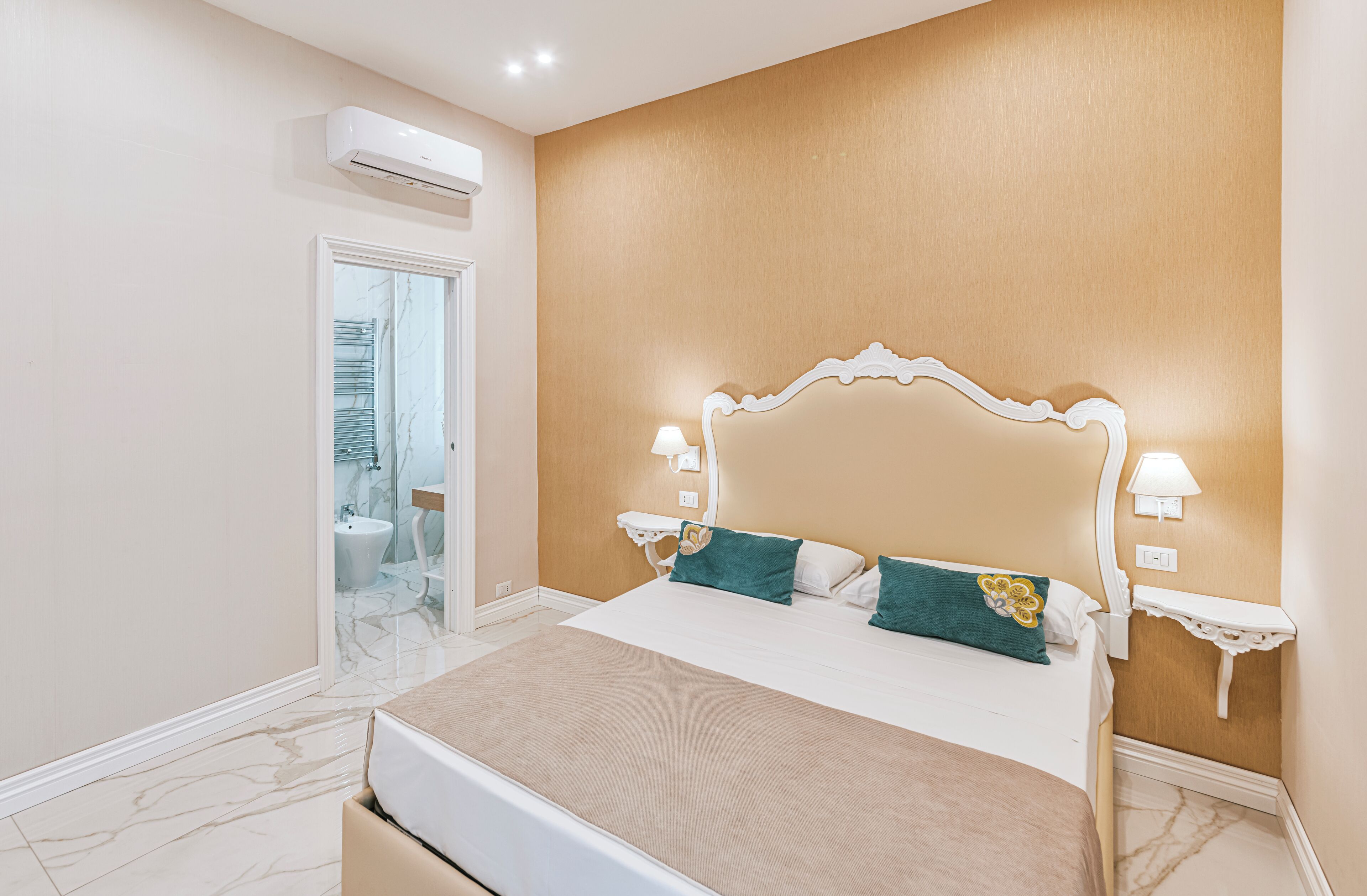 A bedroom with a large bed, two bedside lamps, a nightstand, and a bathroom visible through an open door.