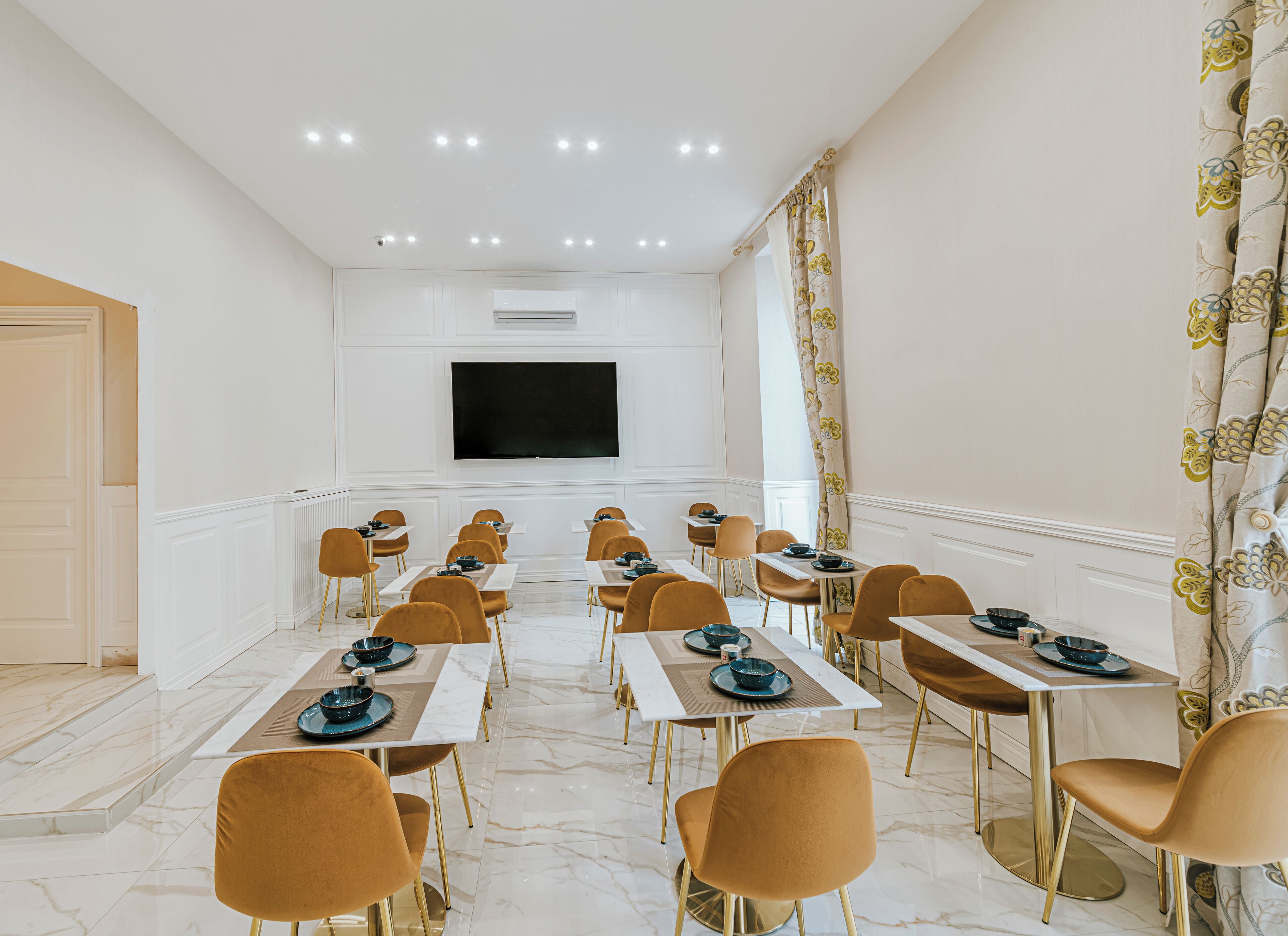 Une salle de conférence aux murs blancs, dotée d’un grand téléviseur à écran plat et de rangées de tables accompagnées de chaises.