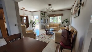 A spacious living room with a large dining table, a sofa, and a coffee table. There are potted plants and a chandelier. - Freeman Greenlees Classical Greek (Rhodes)