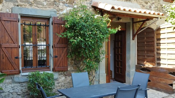 Un bâtiment en pierre avec des volets en bois, un toit en tuiles et une table entourée de chaises devant.
