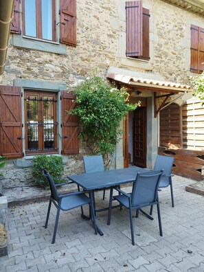 A stone building with wooden shutters, a tiled roof, and a table with chairs in front. - Charmante Maison en Pierres Dans Village, 200m du Canal du Midi. 30 mn Plages (Argeliers)