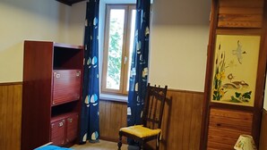 Une chambre avec un lit bleu, une armoire en bois, une chaise, une fenêtre avec des rideaux et un mur orné d’un tableau.