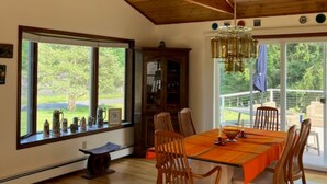 A dining area with a wooden table and chairs, large windows, and a view of greenery outside.