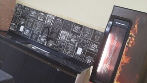 Et køkken med en sort bordplade og et flisebeklædt bagpanel med forskellige kaffemotiver og kagedesign. Et stort Budweiser-køleskab er synligt.