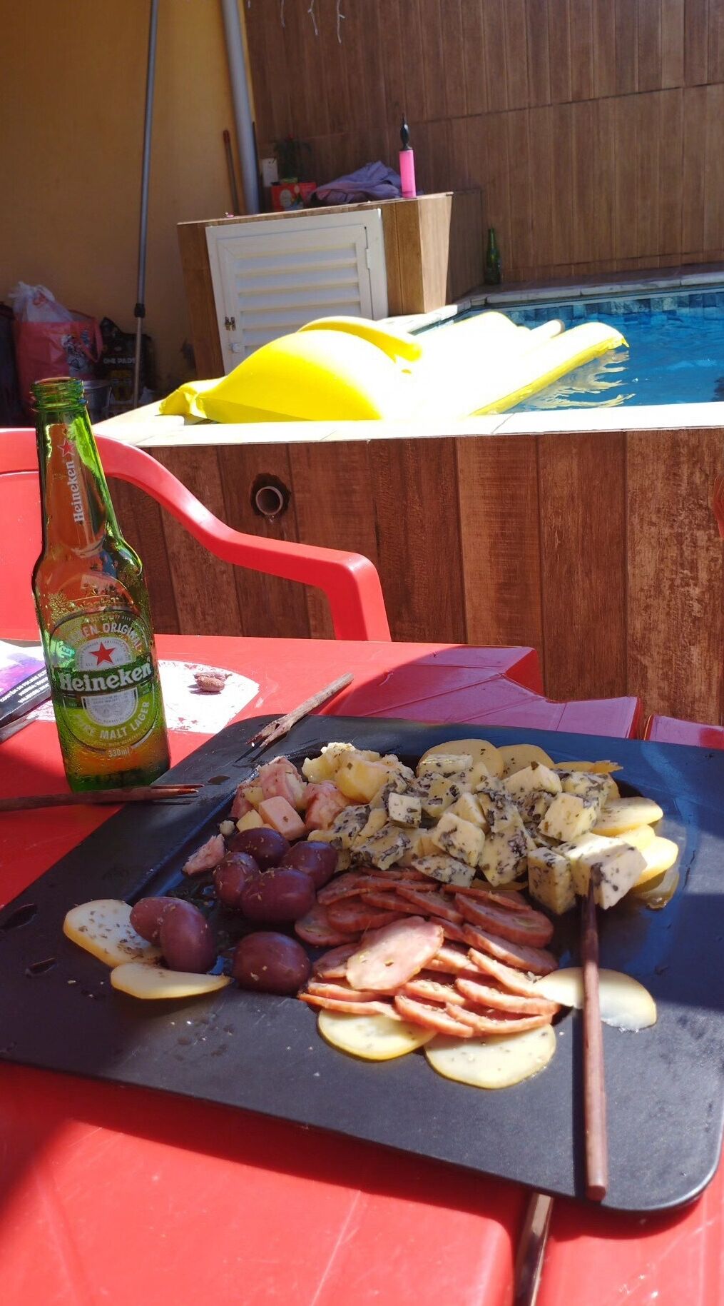 Ein Grillbereich am Pool mit verschiedenen Fleisch- und Käsesorten, einer Heineken-Flasche und einem Pool im Hintergrund.