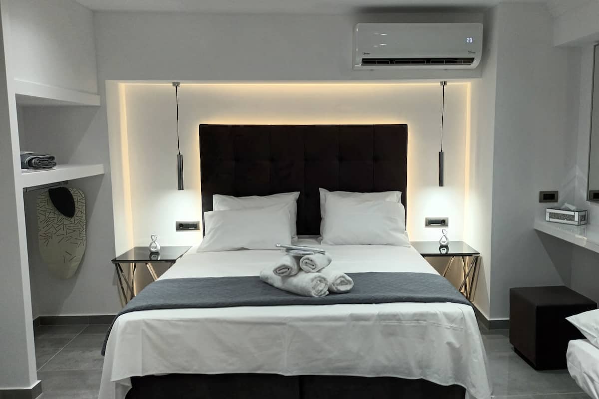 A modern hotel room with a large bed, two bedside tables, a nightstand, a wall-mounted air conditioner, and a small shelf with decorative items.