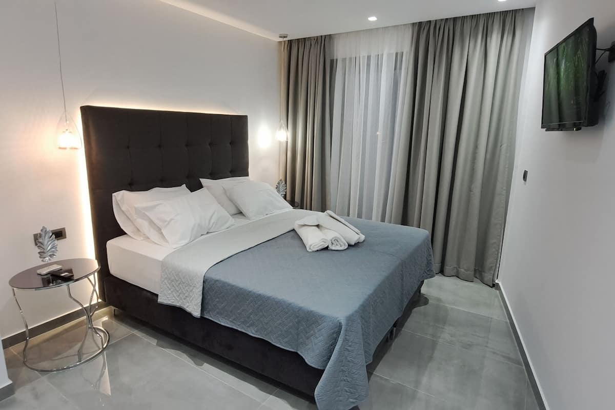 A modern hotel room with a large bed, a bedside table, a flat-screen TV mounted on the wall, and a large window with curtains.