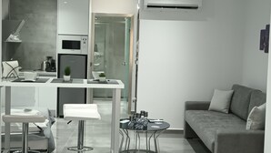 A modern, minimalist living space with a kitchenette, a glass door leading to a bathroom, and a grey sofa. - KM LUXURY apart downtown (Heraklion)