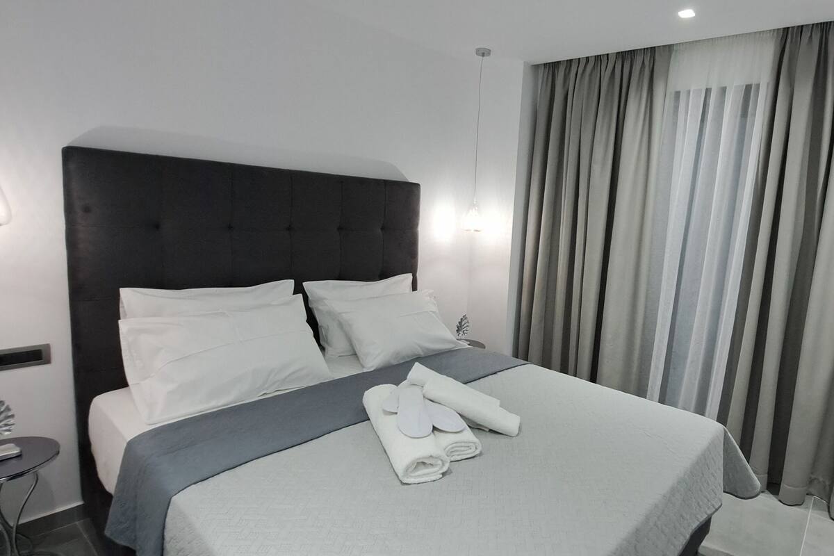 A neatly made bed with white linens, a grey blanket, and a towel with a bathrobe on it. There is a small round table with a remote control on it.