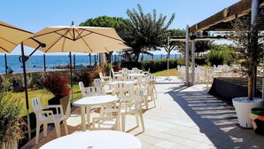 Un patio exterior con mesas y sillas de plástico blanco, sombrillas color beige y vistas al mar.
