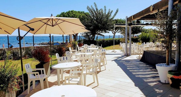 An outdoor patio with white plastic tables and chairs, beige umbrellas, and a view of the sea.