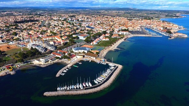 Vista aérea de un pueblo costero con un puerto deportivo, edificios residenciales y un mar azul cristalino.