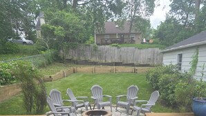 Un patio trasero con un hogar a leña de piedra rodeado de sillas Adirondack, una terraza de madera y una valla de madera.