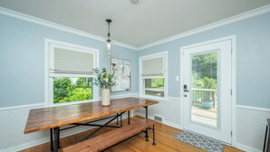 Una habitación con una mesa de madera, dos ventanas con persianas, una puerta con vista y una lámpara colgante.