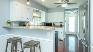 Una cocina con armarios blancos, encimera de granito, dos taburetes de bar, ventilador de techo y refrigerador.