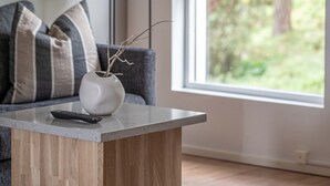 Ein modernes Wohnzimmer mit einem grauen Sofa, einem kleinen Holztisch, einer Vase mit Zweigen und einer Stehlampe. Ein großes Fenster bietet Blick auf Bäume draußen.