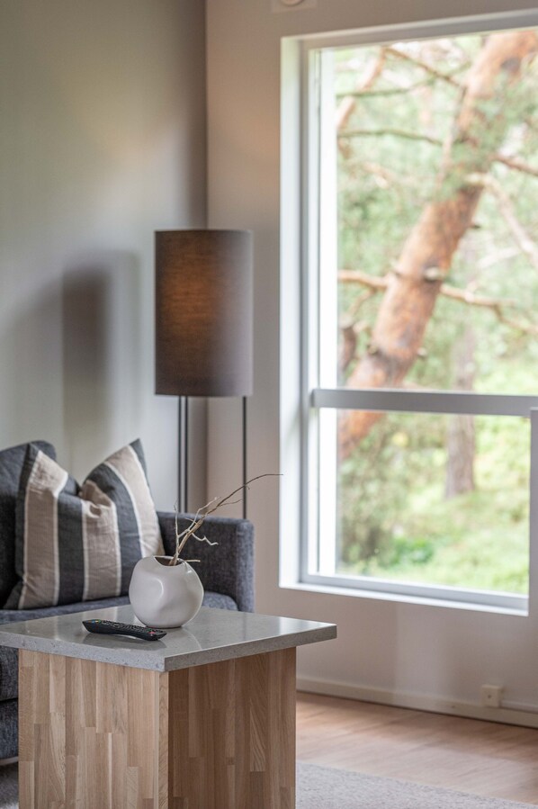 A modern living room with a grey sofa, a small wooden coffee table, a vase with branches, and a floor lamp. A large window offers a view of trees outside. - Tjörnbro Arena (Myggenäs)