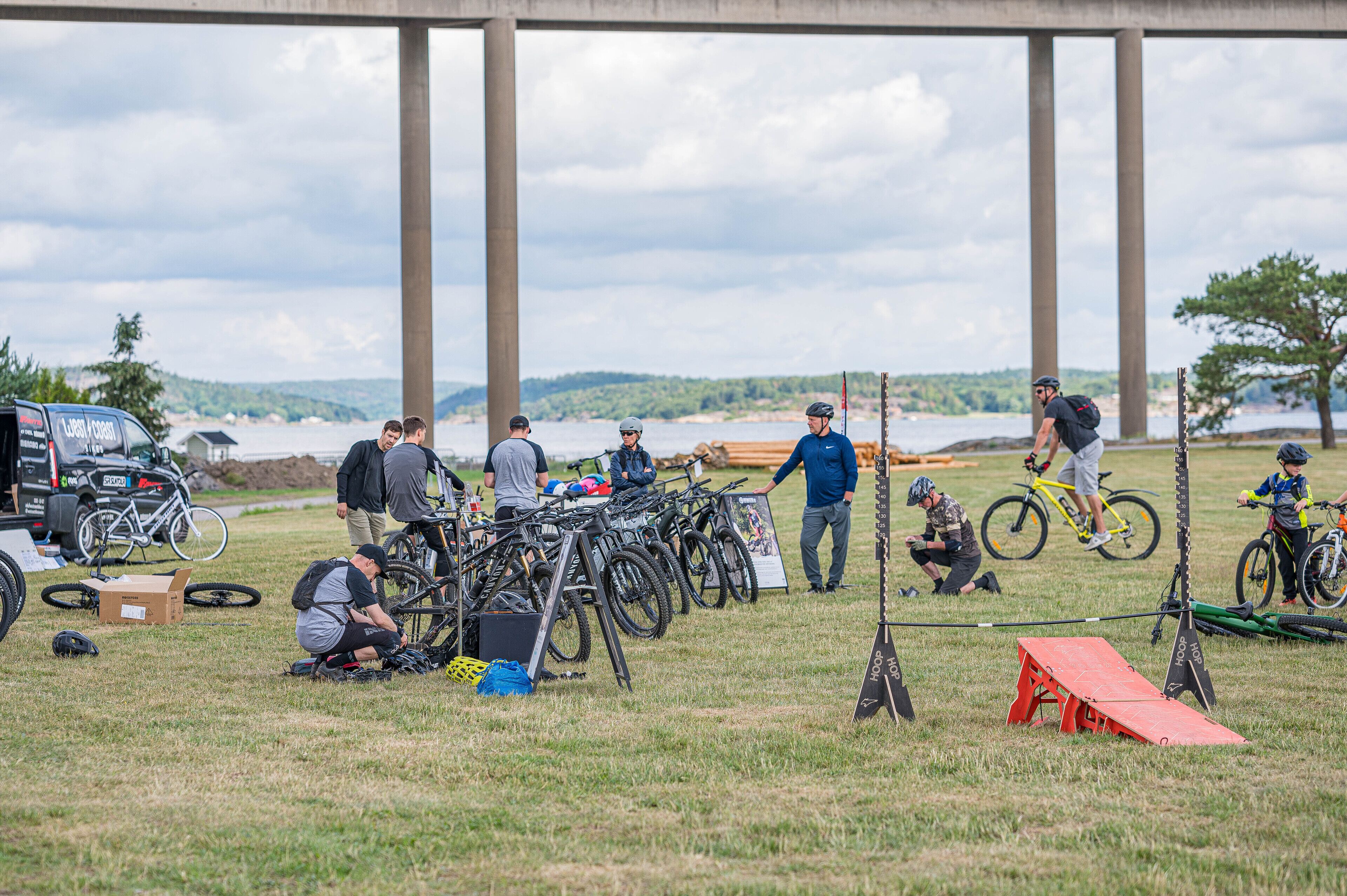 A group of people around bicycles and a truck, with a grassy field and a body of water in the background.