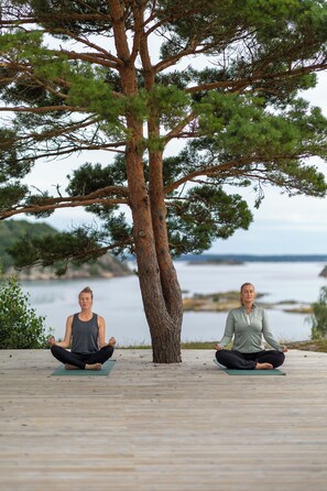 Two people meditating on a wooden deck with a tree and a body of water in the background. - Tjörnbro Arena (Myggenäs)