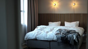 Ein ordentlich bezogenes Bett mit grauem Kopfteil, zwei Wandleuchten und einem mit Vorhängen versehenen Fenster.