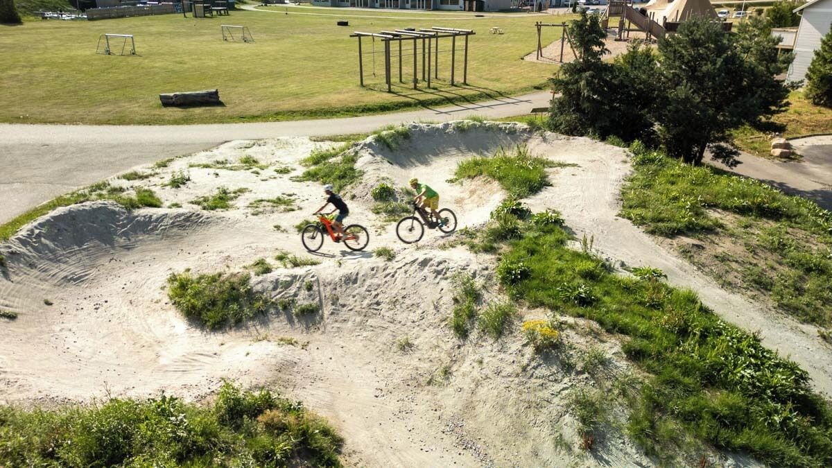 A dirt bike track with two riders, surrounded by grassy areas and a playground.