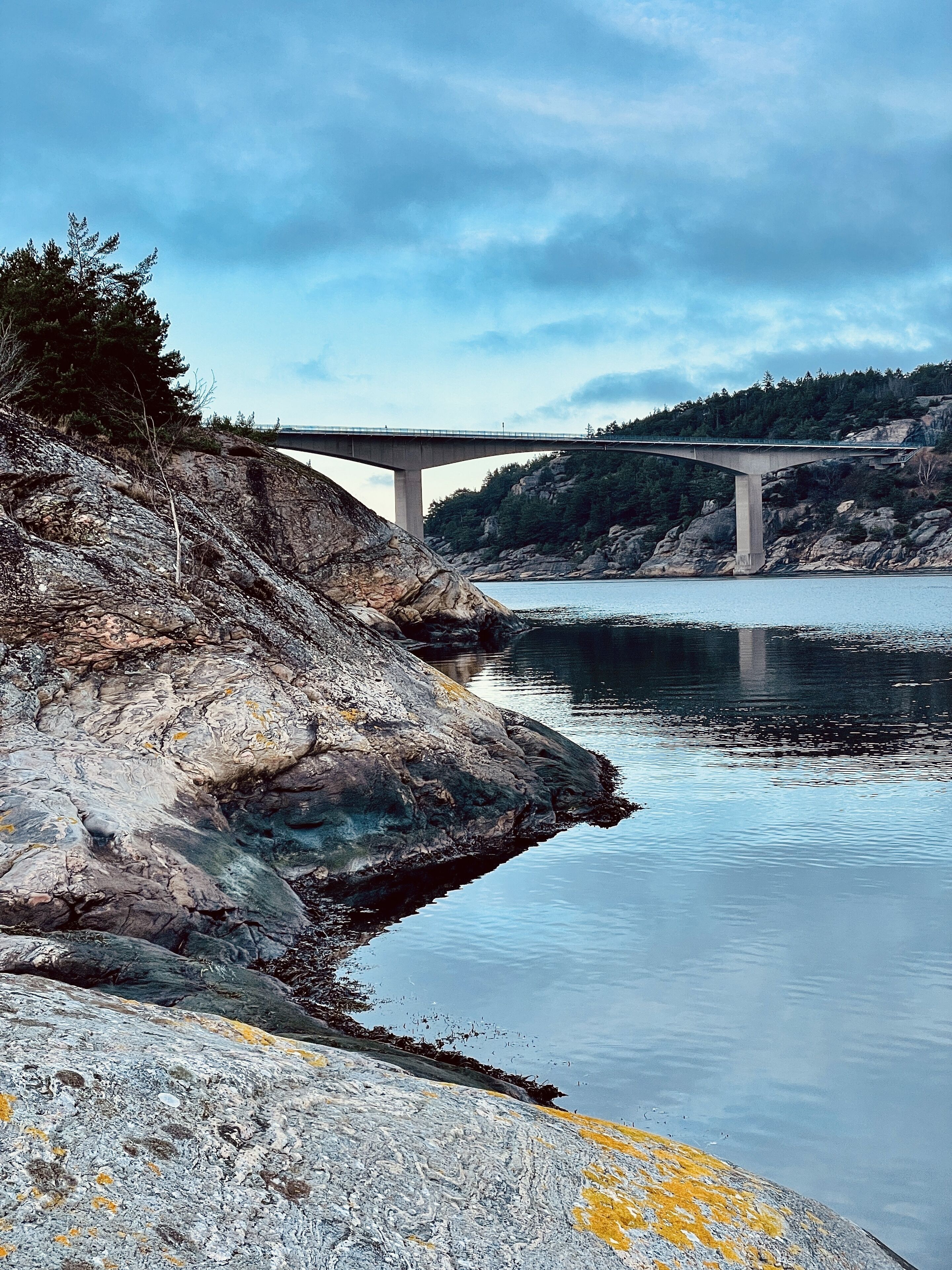 A concrete bridge over a calm river with rocky shores and sparse vegetation.
