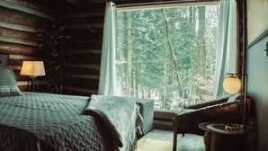 A cozy log cabin bedroom with a large window, a bed, a chair, and a lamp. - Seclude in a Romantic Cabin in an 850 acre Private Forest. (Bernhards Bay)