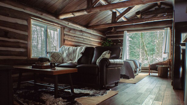 A log cabin interior with a leather sofa, a dining table, and a large window offering a view of trees. - Seclude in a Romantic Cabin in an 850 acre Private Forest. (Bernhards Bay)