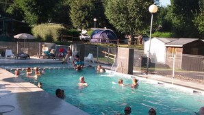 Zona de piscina con varias personas nadando, sillas de descanso y una carpa al fondo.