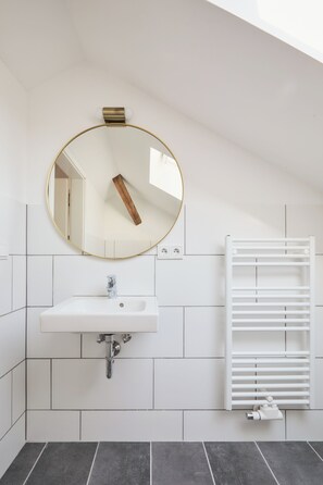 Un baño moderno con un lavamanos blanco, un espejo redondo y un tendedero blanco para toallas.