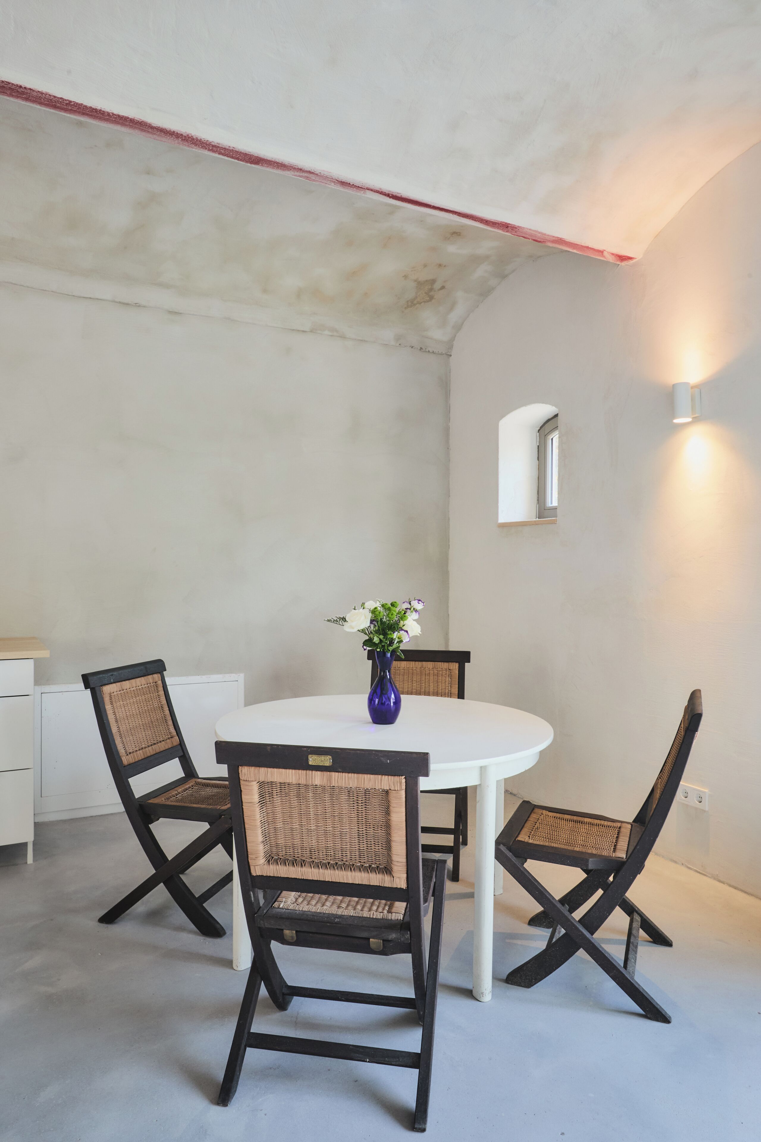 A dining area with a round table, four chairs, a vase with flowers, a wall-mounted light, and a small window.