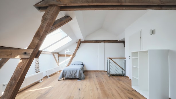 A high-ceilinged attic bedroom with wooden beams, a bed, a white wardrobe, and a staircase.