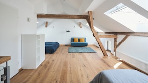 A spacious attic room with wooden beams, a loft bed, a sofa with cushions, and a skylight.