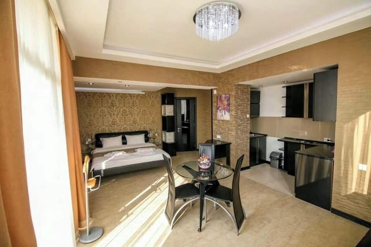 A hotel room with a bed, a dining table with chairs, a small kitchen area, and a chandelier.