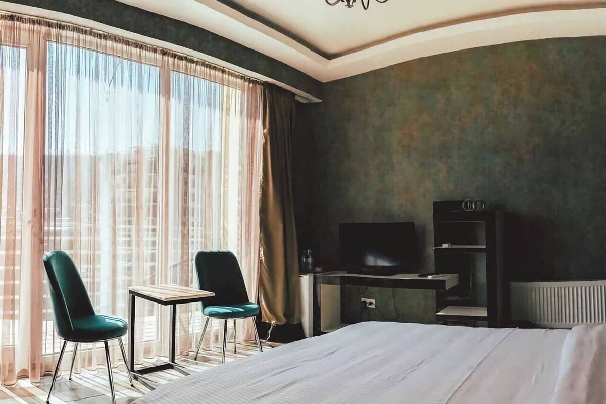 A hotel room with a large bed, two chairs, a desk with a TV, and a window with curtains.