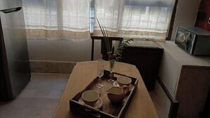 Une petite cuisine équipée d’un réfrigérateur, d’une table sur laquelle se trouve un plateau avec des tasses et un verre, et d’une fenêtre ornée de rideaux légers.