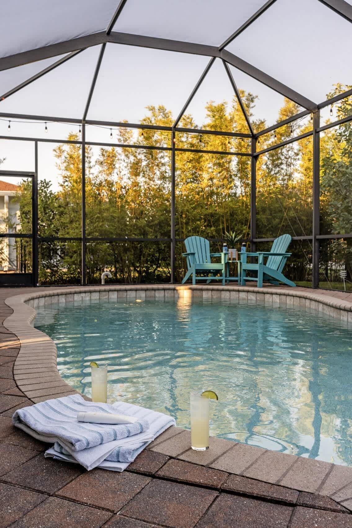An outdoor swimming pool with a covered area, surrounded by trees and a patio with chairs and a table.