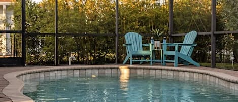 An outdoor swimming pool with a covered area, surrounded by trees and a patio with chairs and a table.