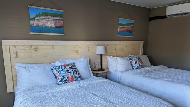 A hotel room with two beds, wooden headboard, white bedding, a nightstand with a lamp, and two wall-mounted paintings. - The Greenland Motel - Room No.2 (Munising)